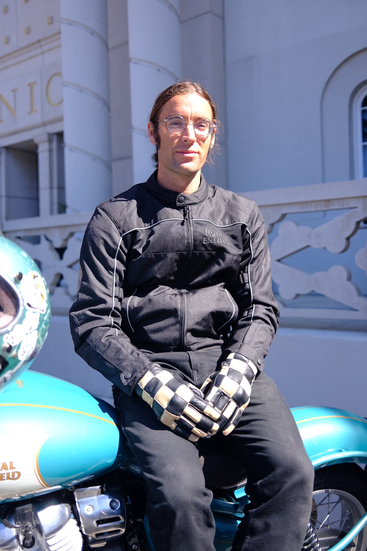 Devin Bagnoli wearing Axel Co checkered gloves sitting on a teal motorcycle with a blurred background